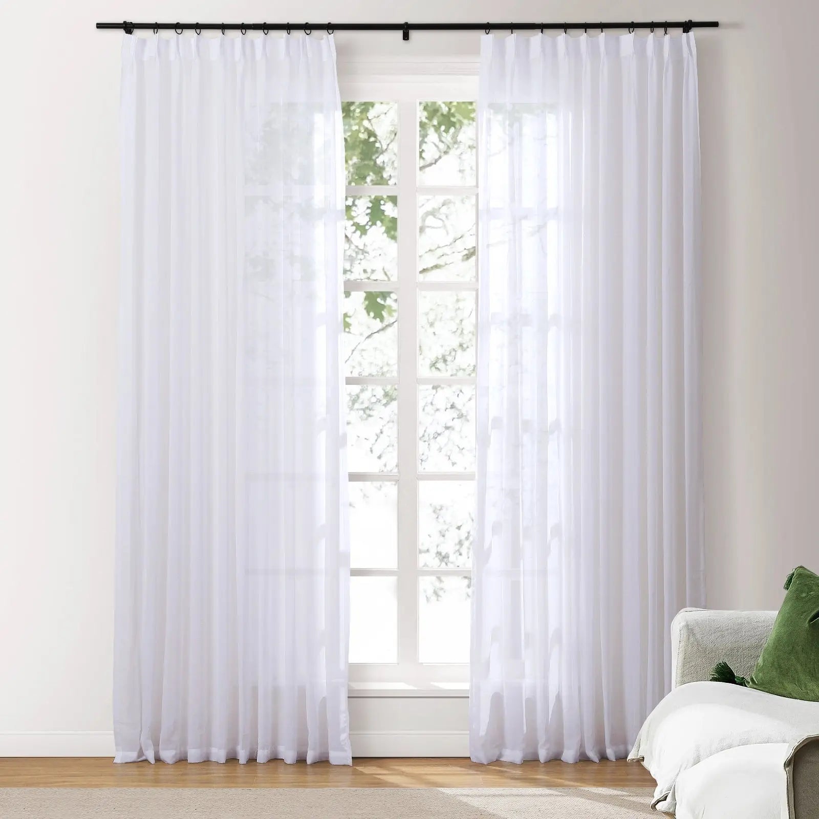 A bright interior room with sheer white rod pocket curtains on a black rod framing a white multi-pane window showing green tree foliage outside beside a light gray sofa with a green decorative pillow