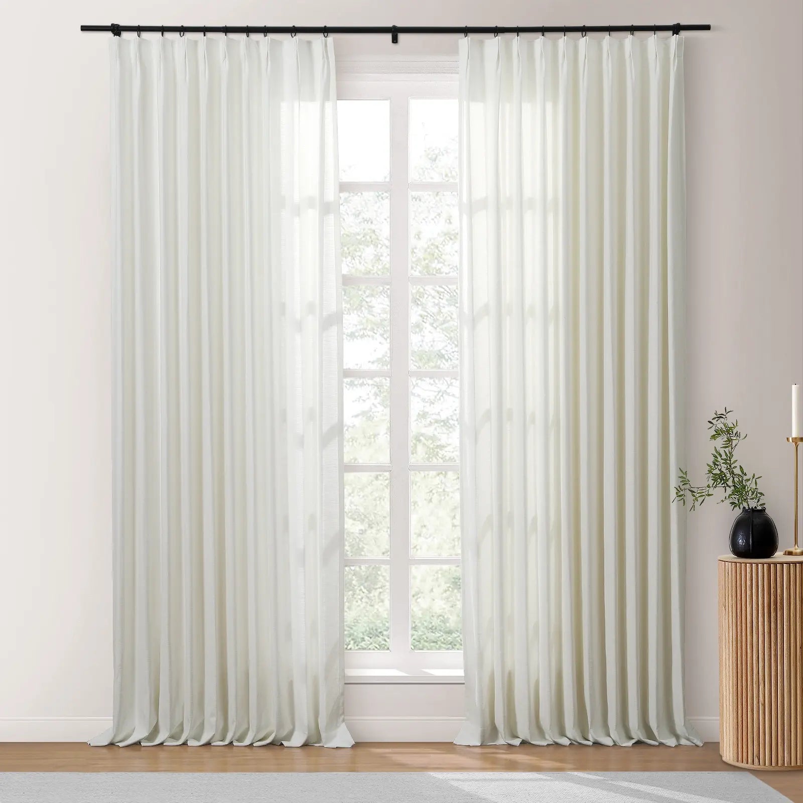 White sheer floor-length pleated curtains hang from a black rod across a large window with a view of green foliage, next to a textured wood side table displaying a black vase with branches and a gold candlestick