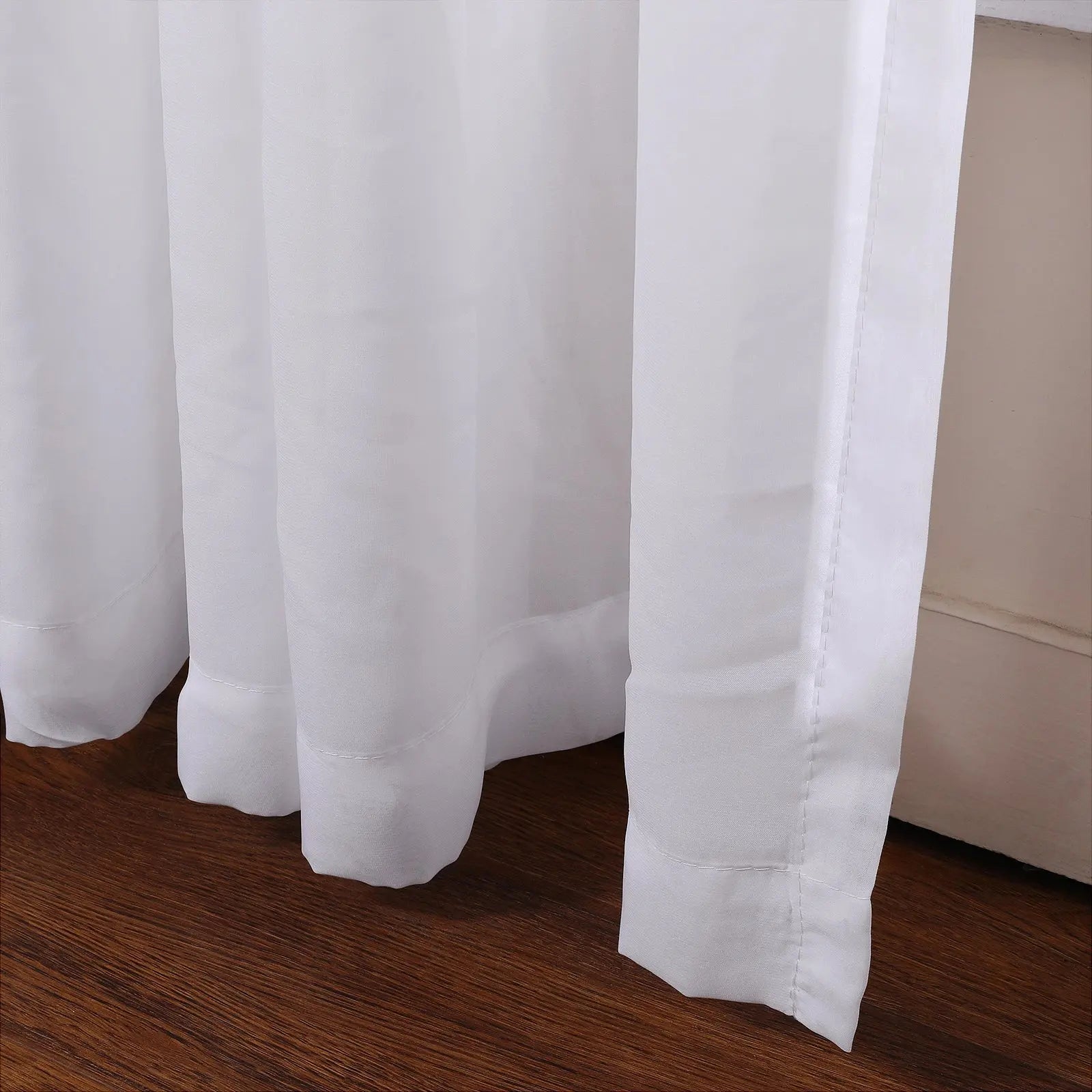 Close-up of flowing white sheer curtains showing soft vertical pleats and neatly hemmed bottoms just above a polished dark wood floor
