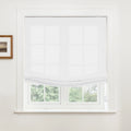 Photograph of a window in an interior room featuring a white semi-sheer Roman shade partially raised revealing lush green trees outside and a small framed landscape print on the adjacent white wall