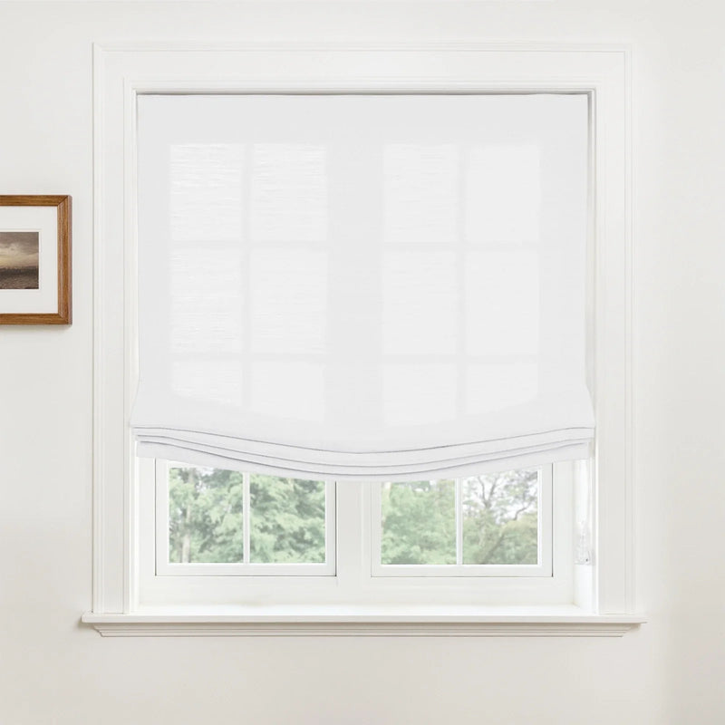 Photograph of a window in an interior room featuring a white semi-sheer Roman shade partially raised revealing lush green trees outside and a small framed landscape print on the adjacent white wall