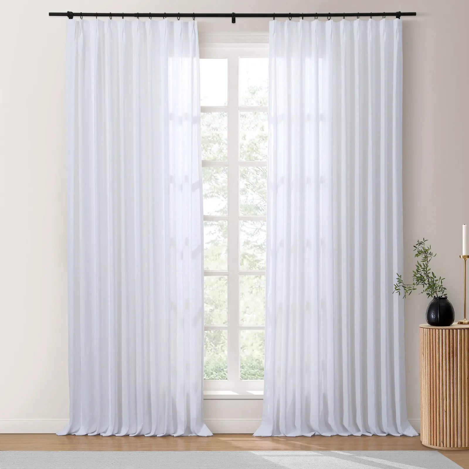 White sheer curtains with subtle vertical stripes hang from a black rod in a bright room, alongside a fluted wooden side table and a black vase with green foliage