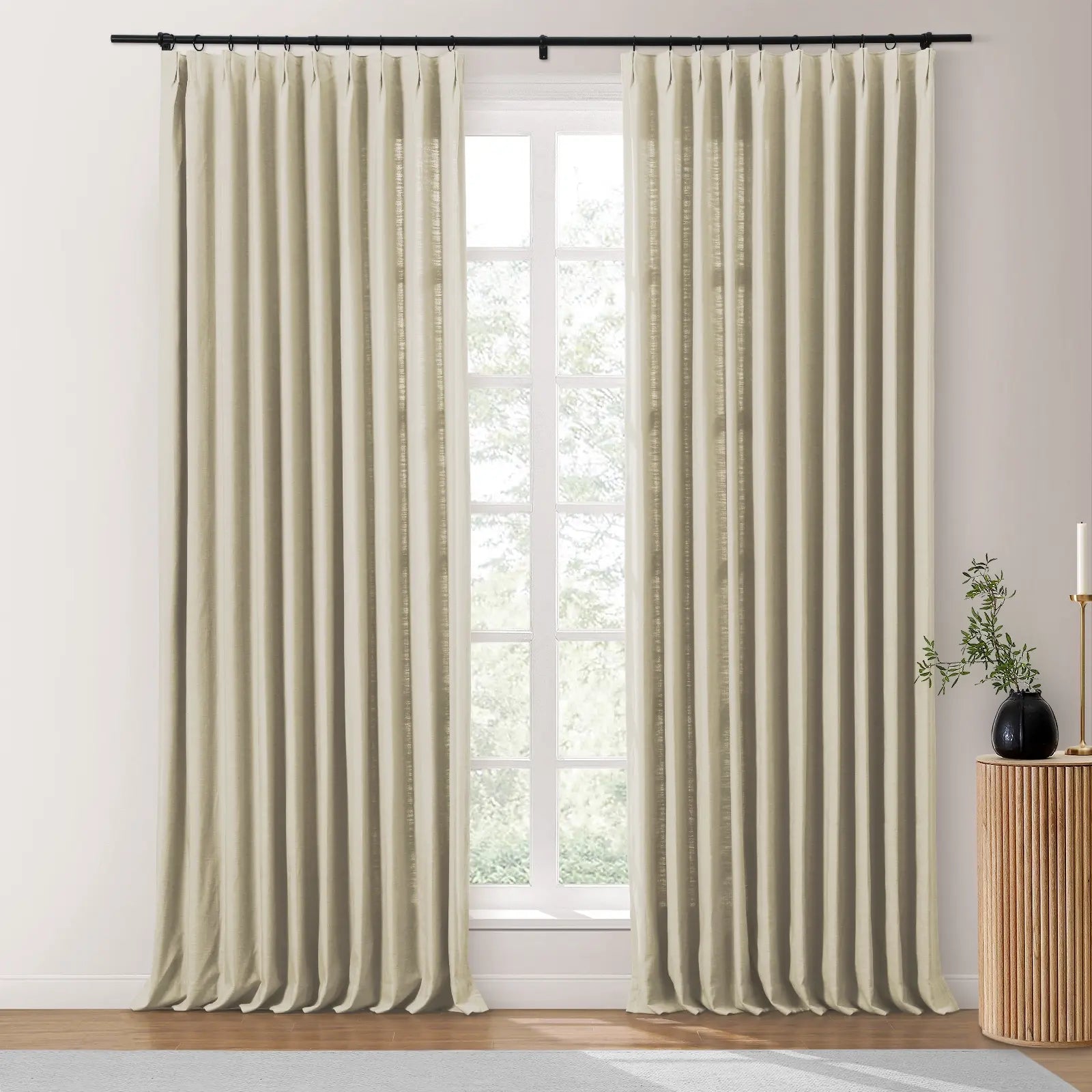 Beige linen pinch-pleat curtains hang from a black rod in a bright contemporary room with a large window revealing green foliage, next to a fluted wooden side table topped with a black vase of branches and a gold candlestick