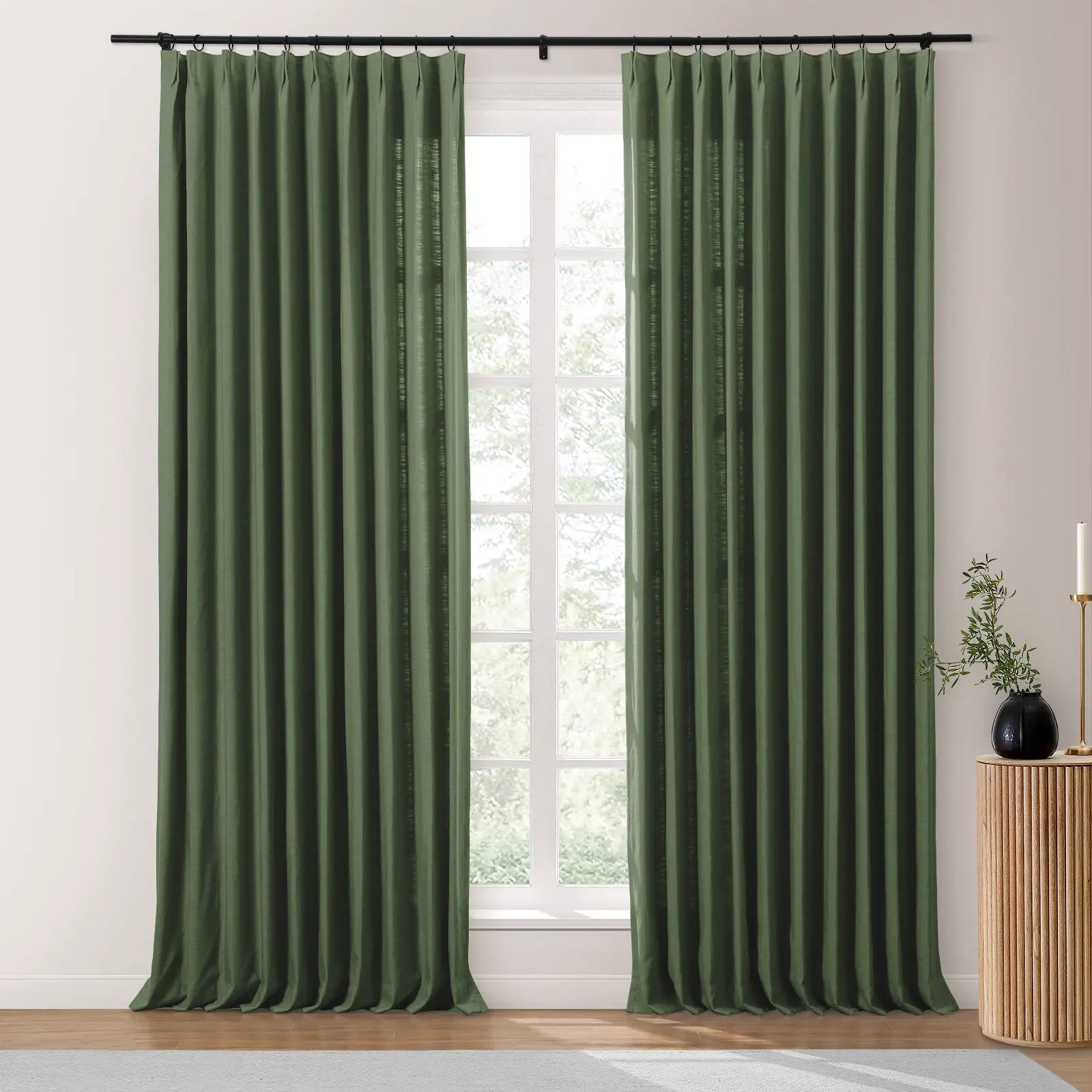 Two dark green pleated linen curtains hang from a black rod flanking a bright multi-pane window showing blurred outdoor greenery, beside a fluted wooden side table topped with a black vase holding green sprigs and a gold candle holder