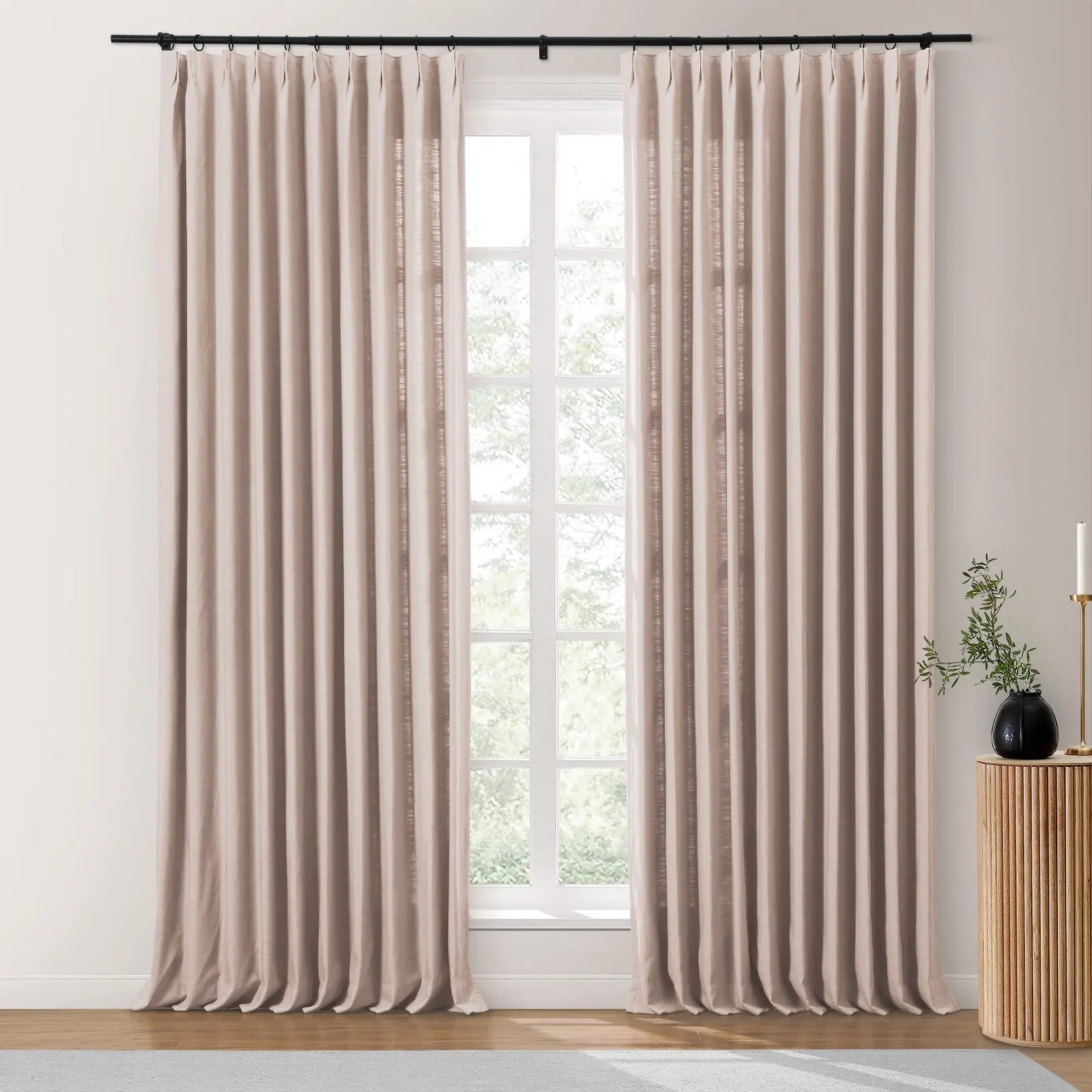 Photograph of two pleated beige linen-blend curtains on a black rod framing a window with a view of green trees, beside a ribbed wooden side table displaying a black vase with branches and a gold candlestick