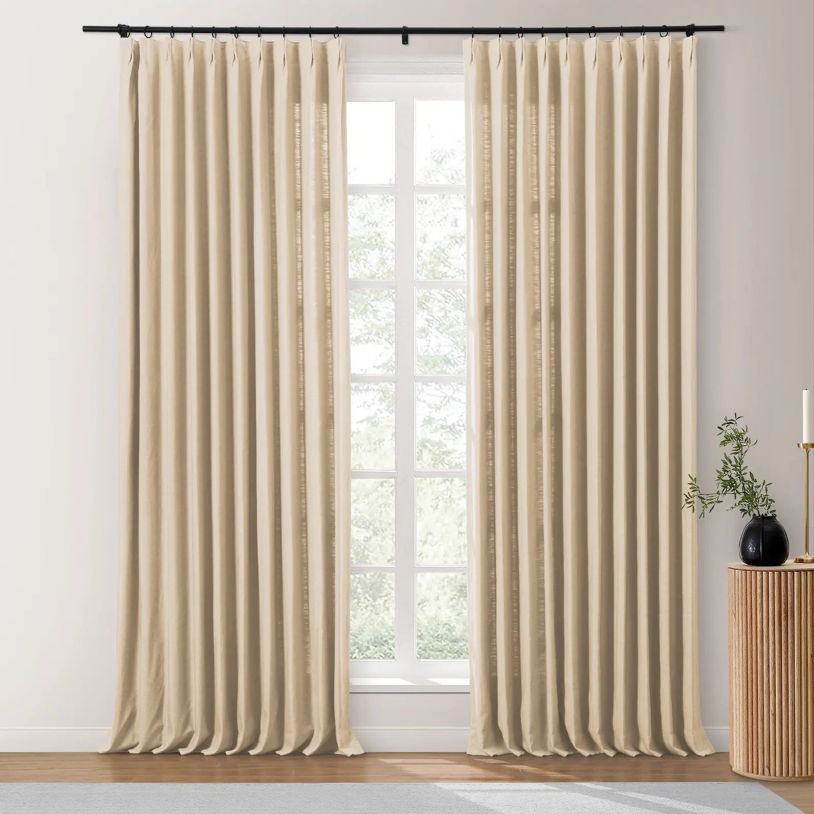 Neutral beige pinch pleat window curtains hung from a black rod in a bright room featuring a fluted wood side table with a dark vase and green plant