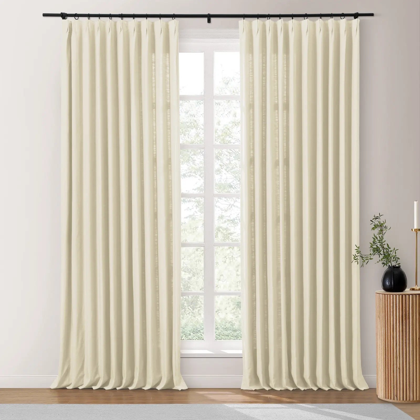 Cream pinch-pleated drapes in a natural textured fabric hang from a black rod, framing a bright window and styled with a fluted wooden side table holding a dark vase with green leaves