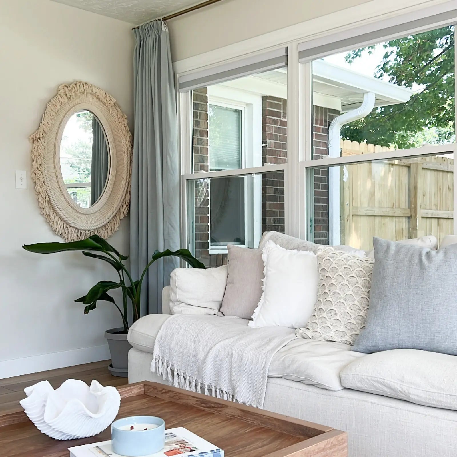 A cozy living room interior with a white linen sofa adorned with cream and light blue textured pillows, a large boho macrame oval mirror on the wall, and expansive windows offering a view of a brick house and wooden fence