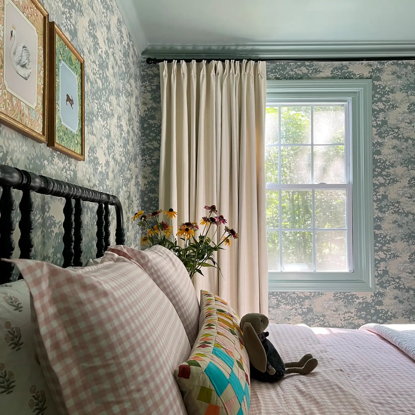 Photograph of a cozy bedroom interior with pink gingham bedding, a black headboard, a gray stuffed bunny, and a colorful quilted pillow, with a bouquet of yellow and purple coneflowers beside cream drapes, against blue and white botanical wallpaper and light blue window trim