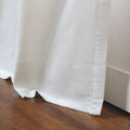 A close-up of white, slightly textured fabric with soft folds resting on a dark brown polished wooden floor beside a white baseboard and a visible side hem