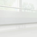 Photograph of a modern white day-night roller blind featuring alternating sheer and opaque horizontal fabric strips, a translucent plastic pull handle at the bottom, and blurred green foliage visible through the window