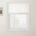 Photograph of a bright room featuring a window with a white frame and a half-drawn off-white linen roman blind above a blurred view of trees, with a framed Van Gogh print on the light beige wall to the left