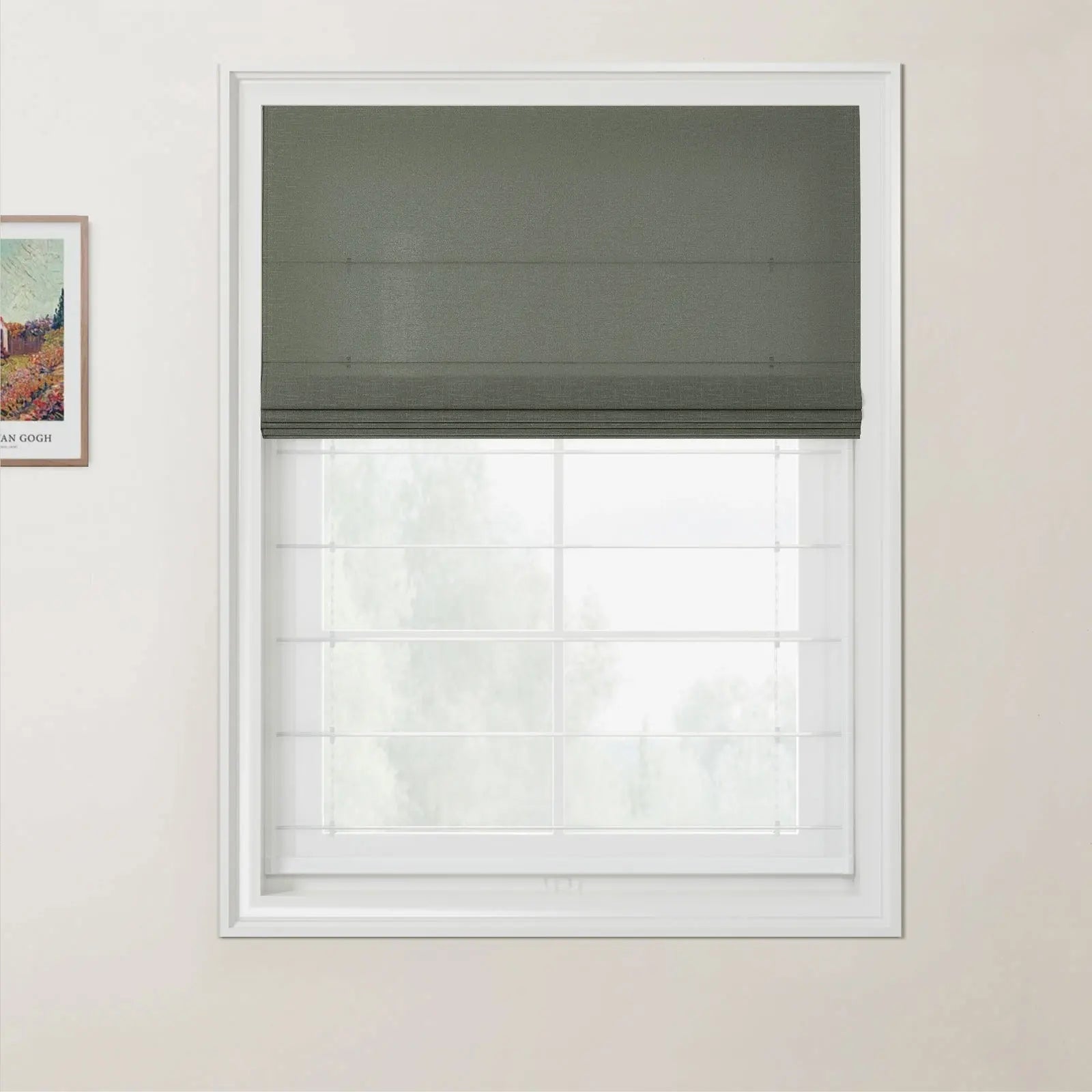 A white framed window with a dark green textured Roman blind partially lowered over a bright view of outdoor trees with a framed Van Gogh print visible on the light beige wall