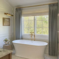 A bright luxury bathroom featuring a white freestanding tub with elegant brass fixtures, flanked by pale blue velvet curtains on a brass rod and a large window showcasing vibrant green trees, detailed with classic wainscoting and a small framed landscape artwork