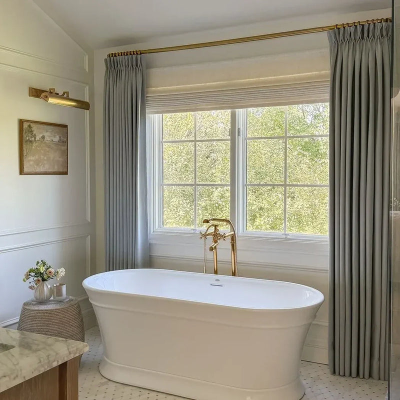A bright luxury bathroom featuring a white freestanding tub with elegant brass fixtures, flanked by pale blue velvet curtains on a brass rod and a large window showcasing vibrant green trees, detailed with classic wainscoting and a small framed landscape artwork