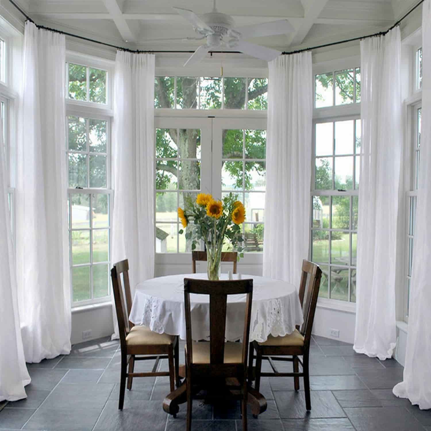 A bright sunroom featuring large bay windows with sheer white curtains a round table adorned with a white lace tablecloth and vibrant yellow sunflowers in a vase all set on a dark slate tile floor