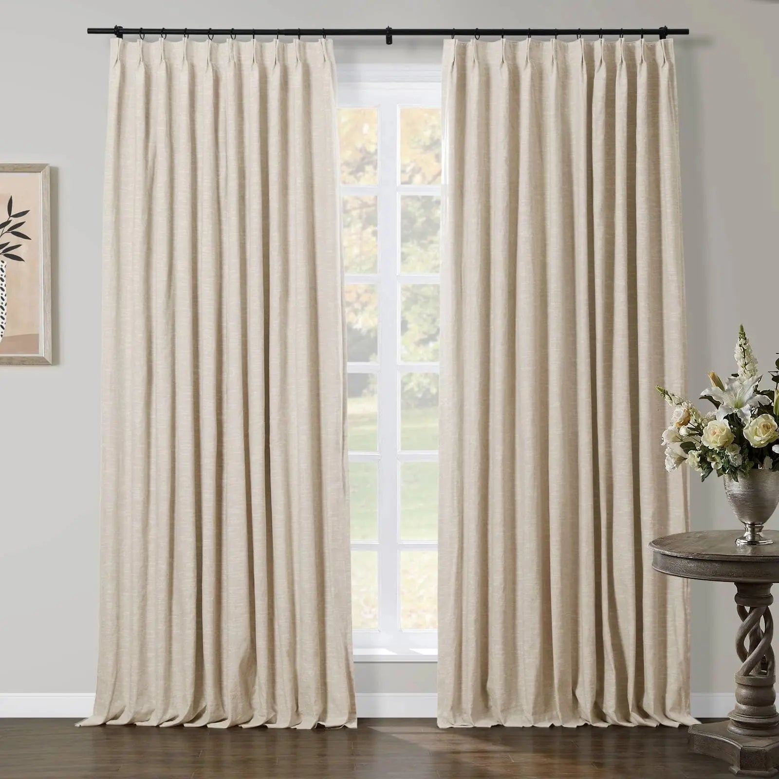 Light beige pleated curtains on a black rod frame a large window with a soft green outdoor view in an elegant room, featuring a twisted dark wood side table with a silver vase of white and cream flowers