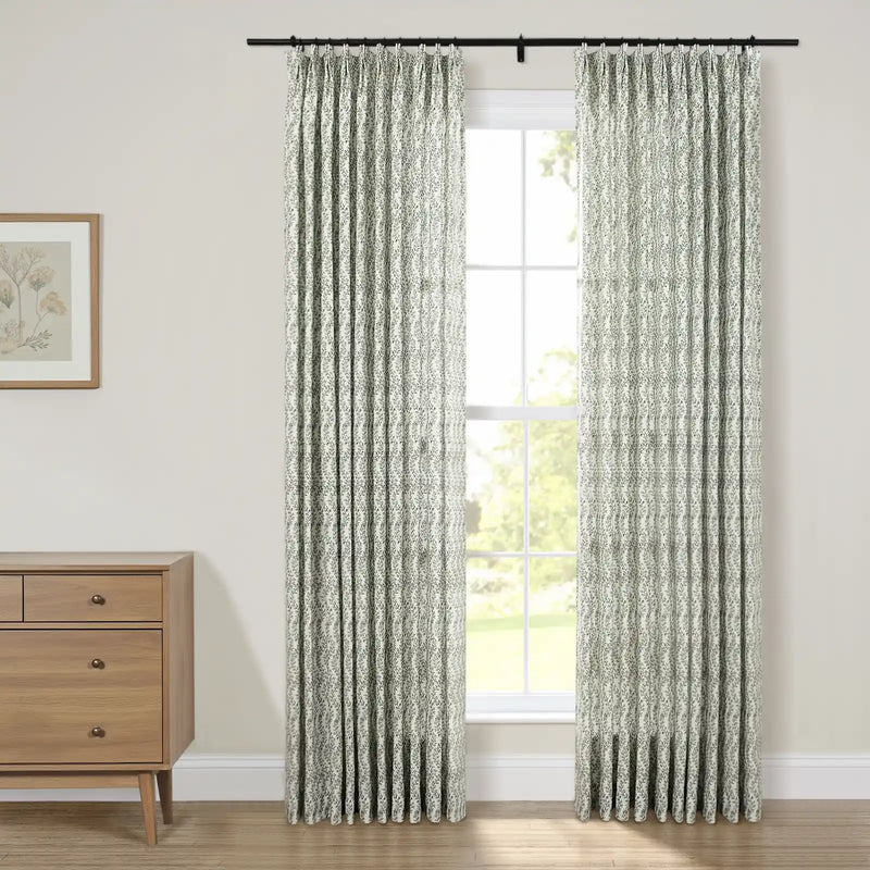 Photograph of a modern room interior featuring a window with green leaf patterned white curtains on a black rod, adjacent to a light wood mid-century modern dresser with a framed botanical print
