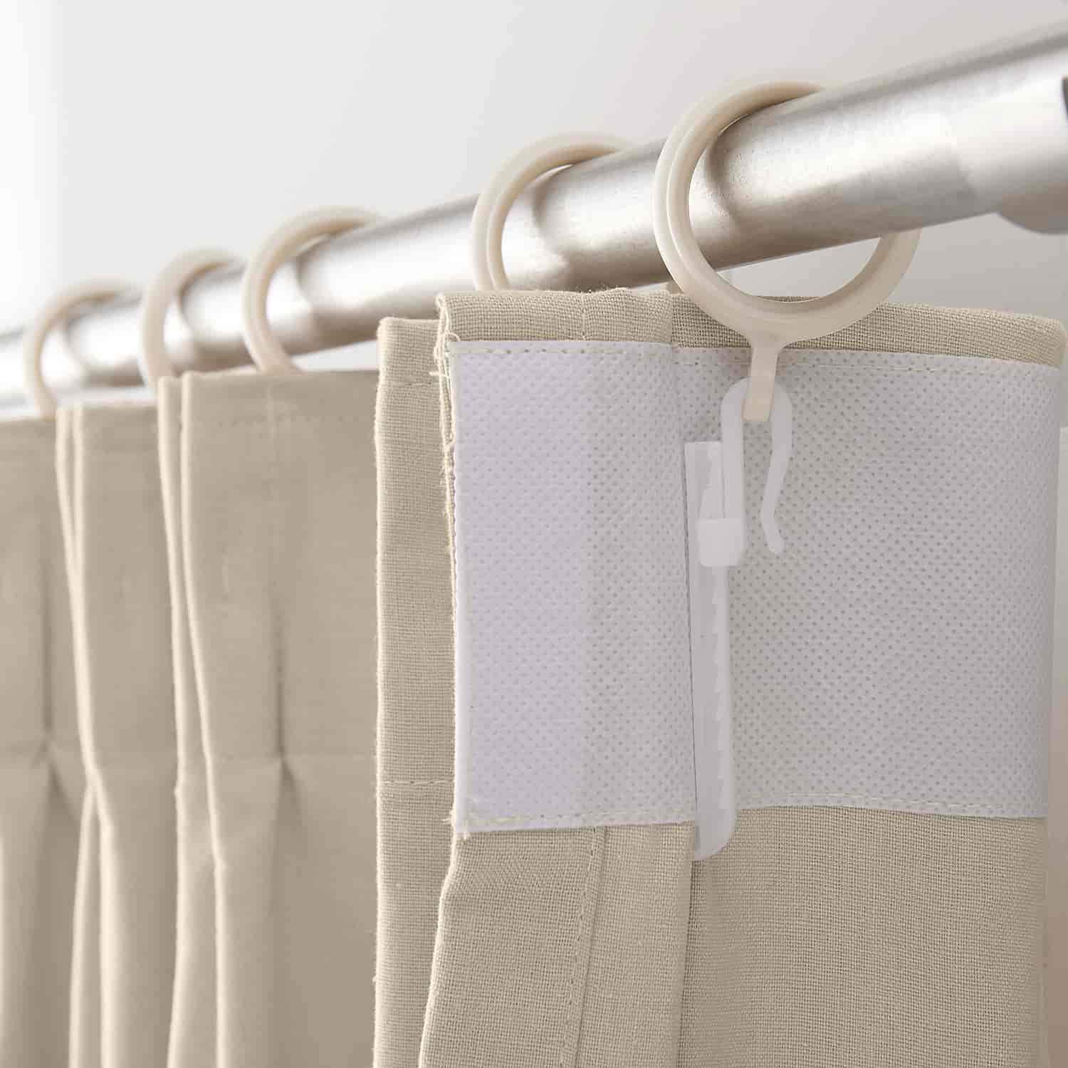 Beige pleated curtains with a linen-like texture hanging from a silver rod with white plastic rings and a visible white non-woven reinforcing strip