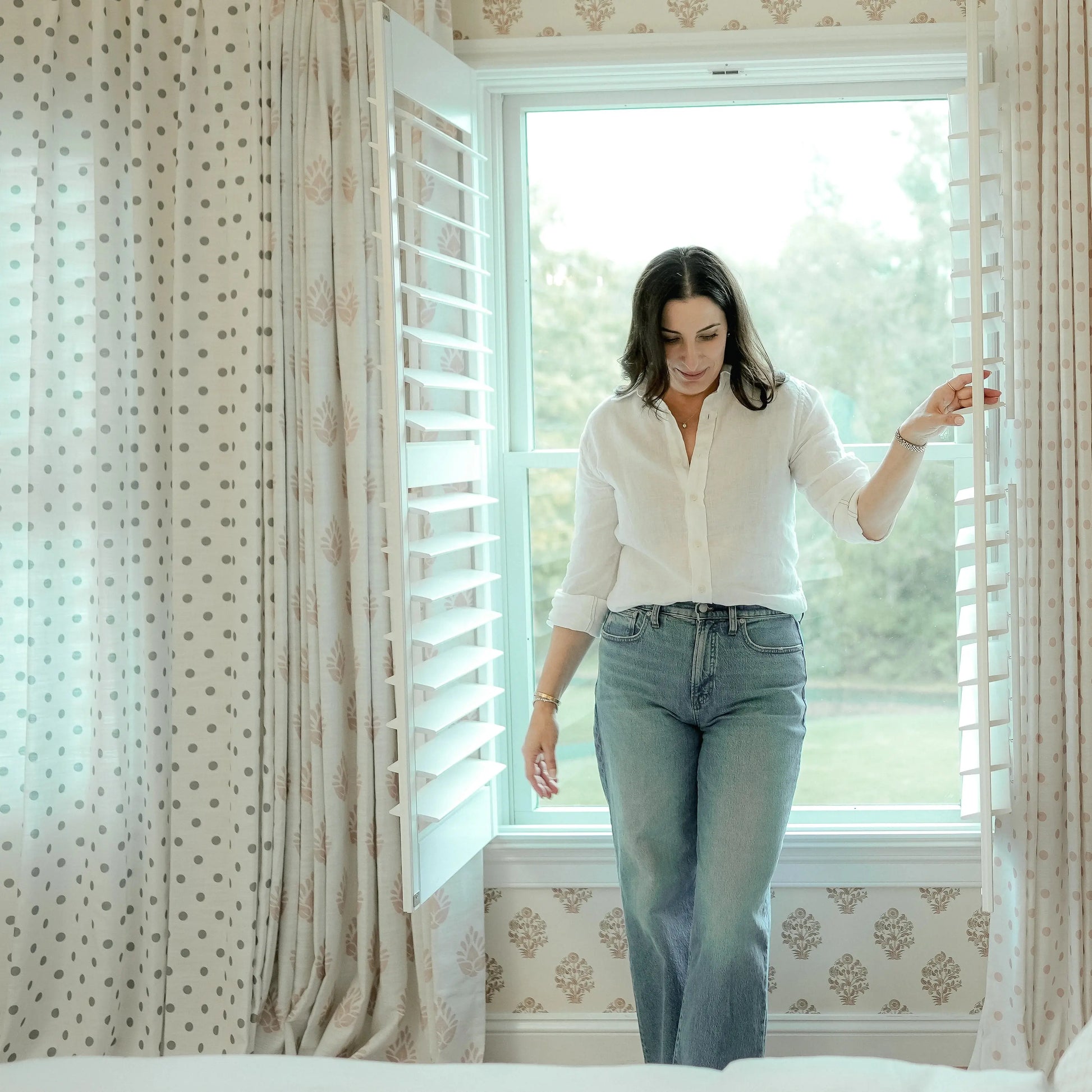 A woman in a white linen shirt and jeans stands by a window, adjusting a white louvered shutter next to gray polka dot curtains and beige floral wallpaper