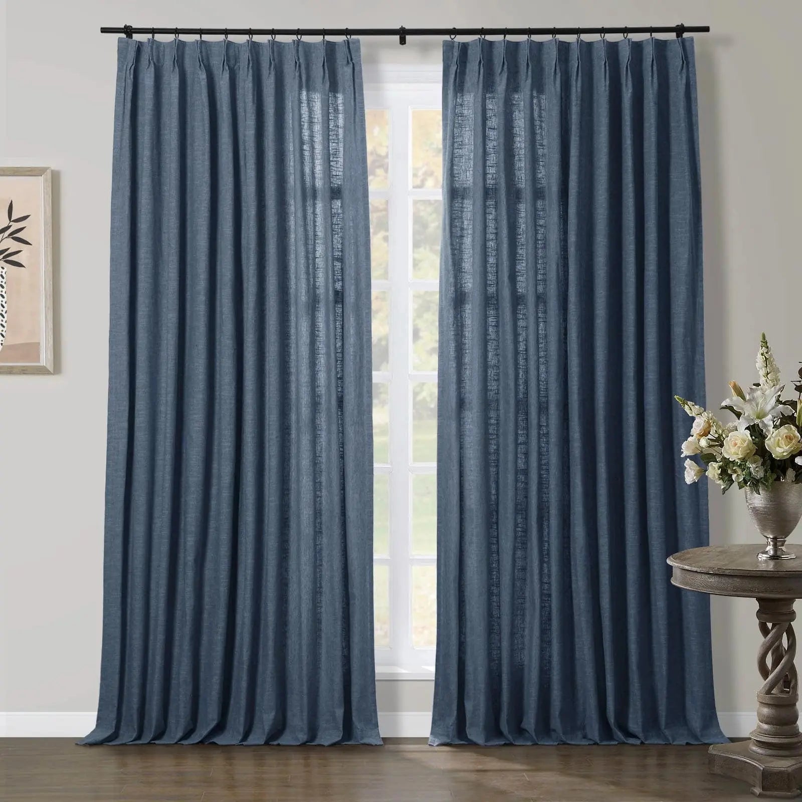Blue linen pinch pleat curtains in an interior room with a large window, displaying a deep denim hue, a subtle textured weave, and pooling elegantly on a dark wooden floor, alongside a white floral arrangement