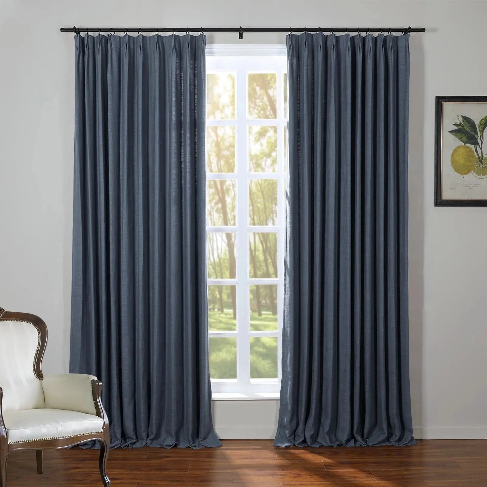 A home interior featuring dark blue linen curtains on a black rod framing a white multi-pane window with bright sunlit trees outside, a cream upholstered armchair with wooden trim is on the left, and a framed botanical lemon print hangs on the right wall above a dark wood floor