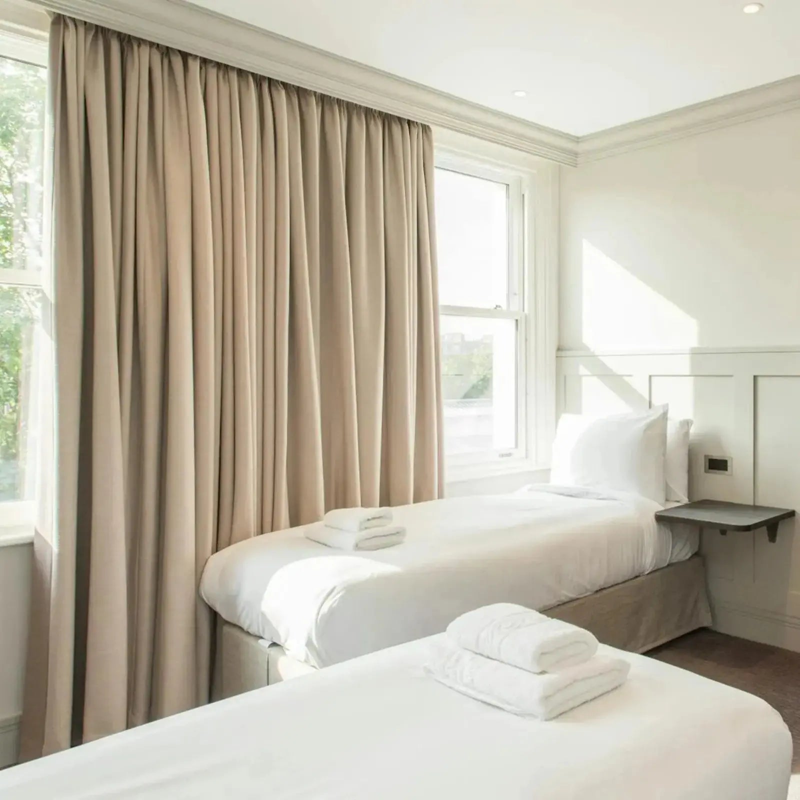 A bright minimalist twin hotel room with two beds featuring crisp white bedding and neatly folded white towels floor-length taupe curtains at a large window and sunbeams illuminating a dark floating nightstand