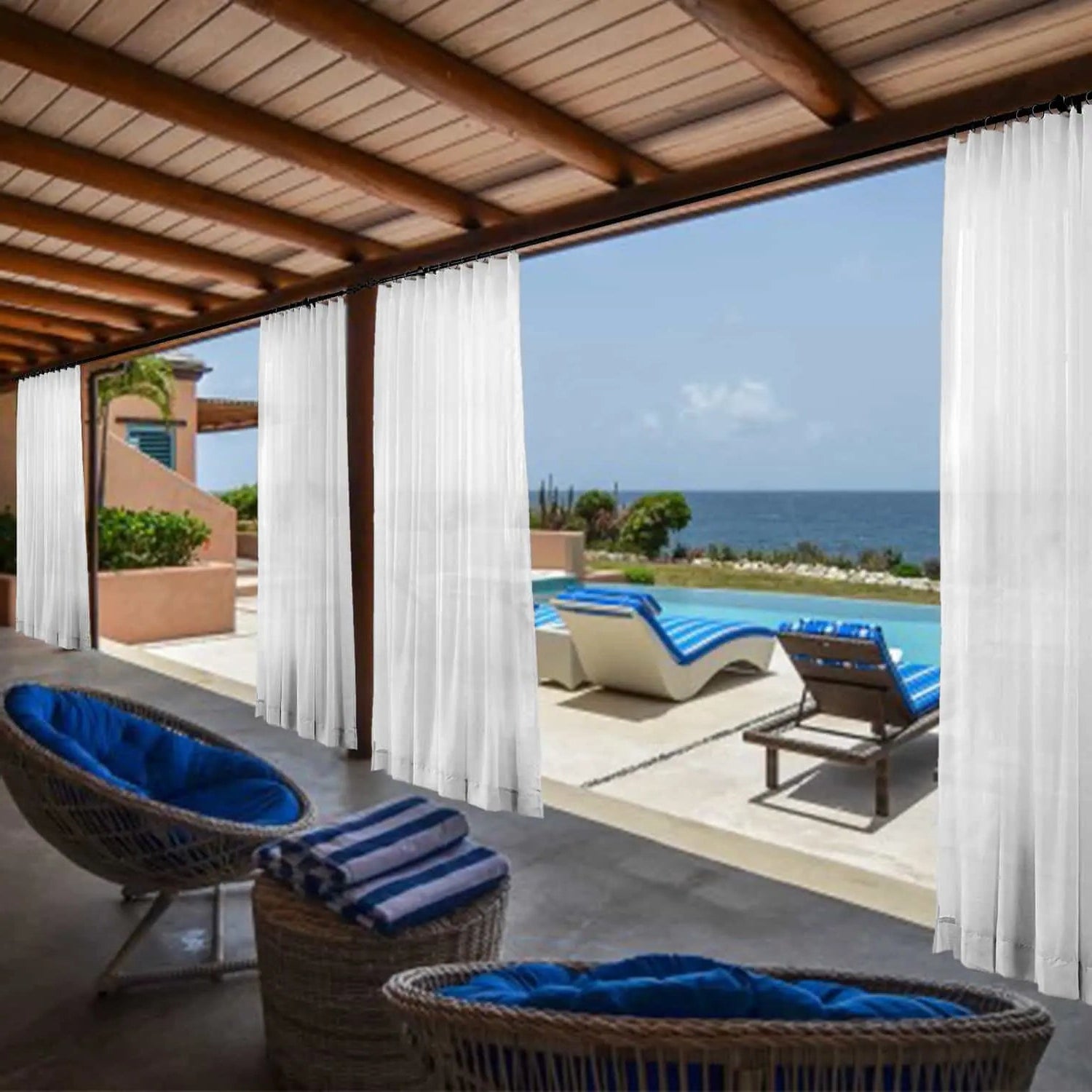 A tranquil outdoor veranda with a wooden pergola and flowing white sheer curtains features woven patio furniture with vibrant blue cushions and striped towels, all overlooking an infinity pool with matching blue-striped lounge chairs and the clear blue ocean