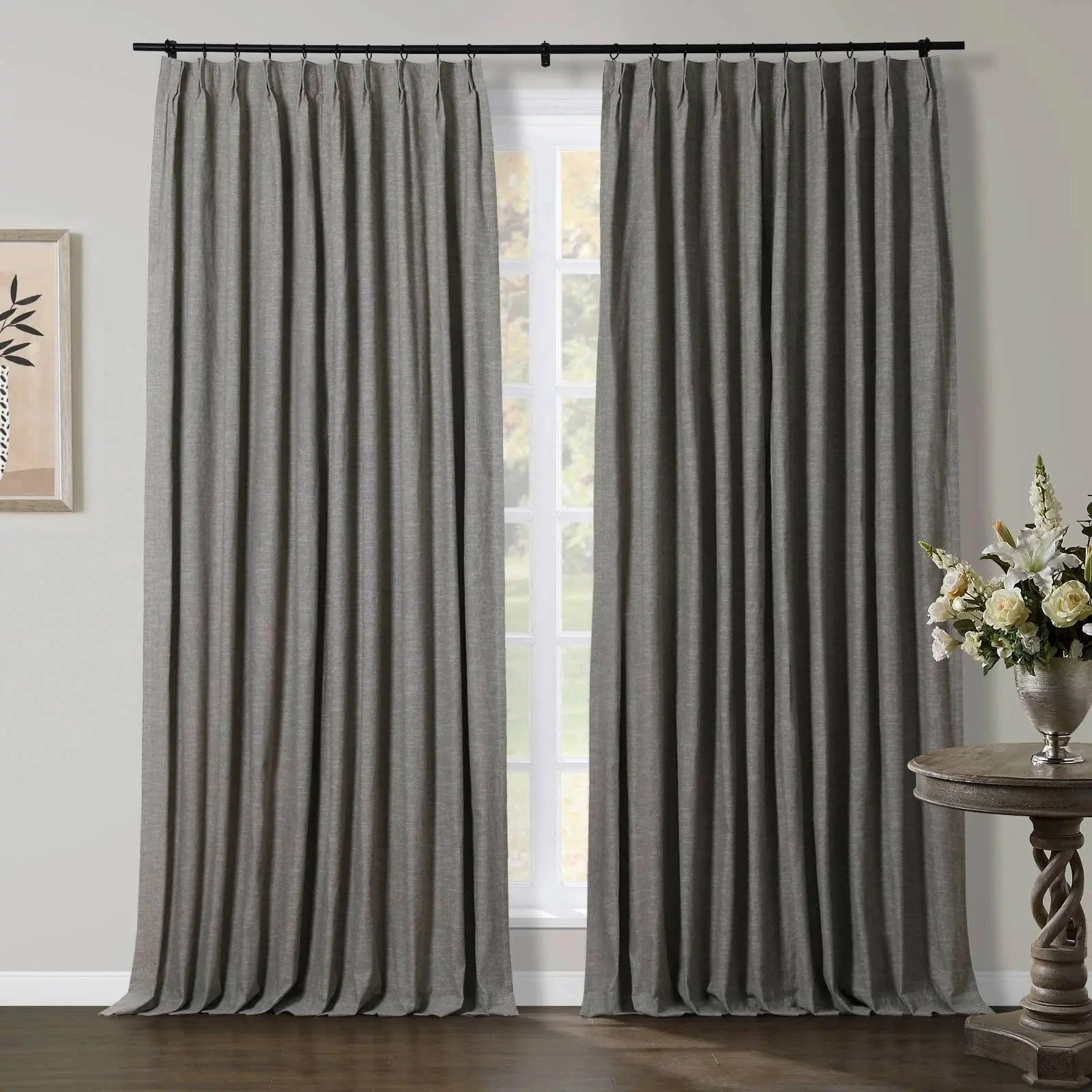 Grey pleated curtains hang from a black rod over a window, flanked by abstract wall art and a silver vase with a white floral arrangement on a carved wooden table