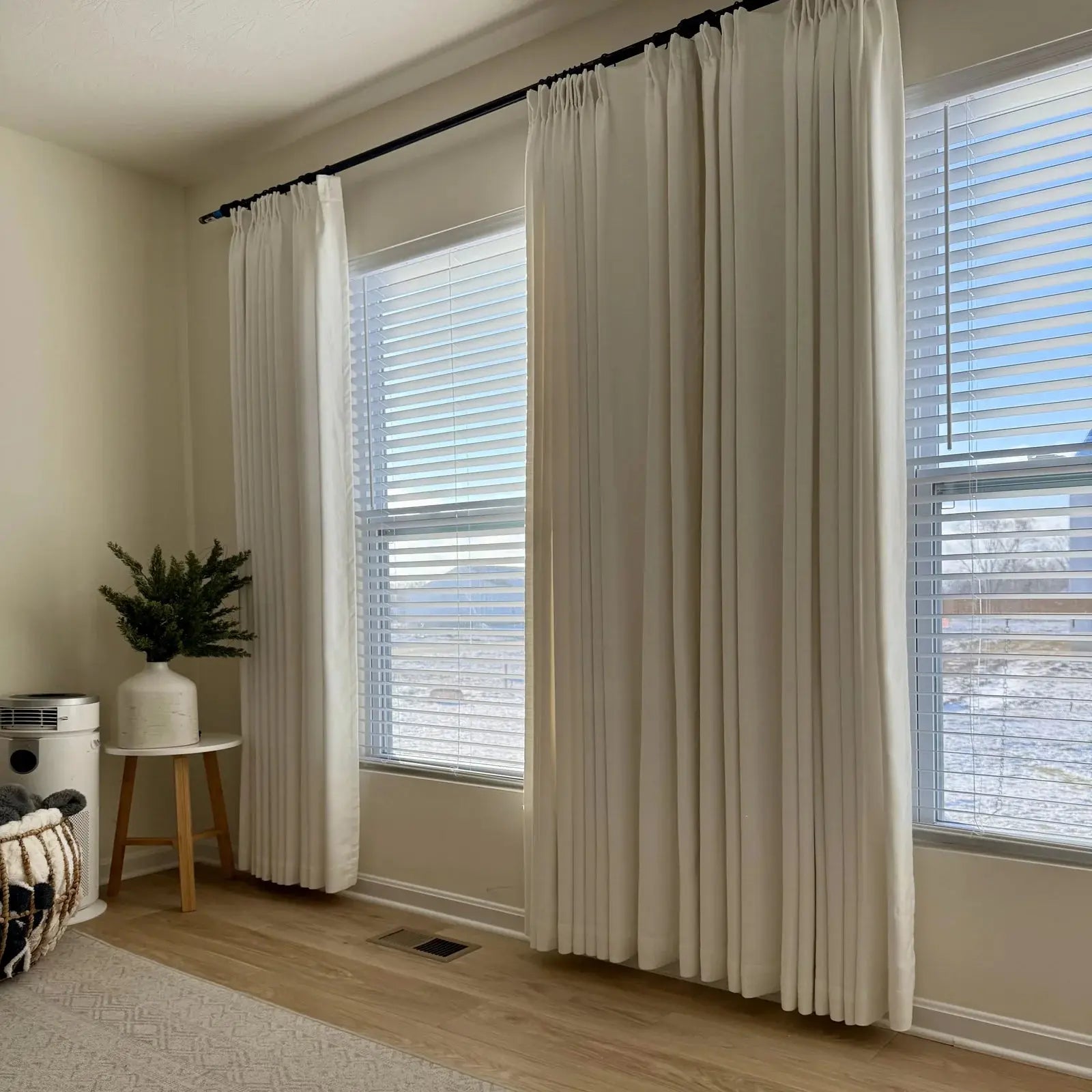 Interior view of a brightly lit room showcasing two windows with flowing cream-colored curtains on black rods and white Venetian blinds, beside a small side table holding a green plant in a white vase, looking out onto a snowy winter landscape