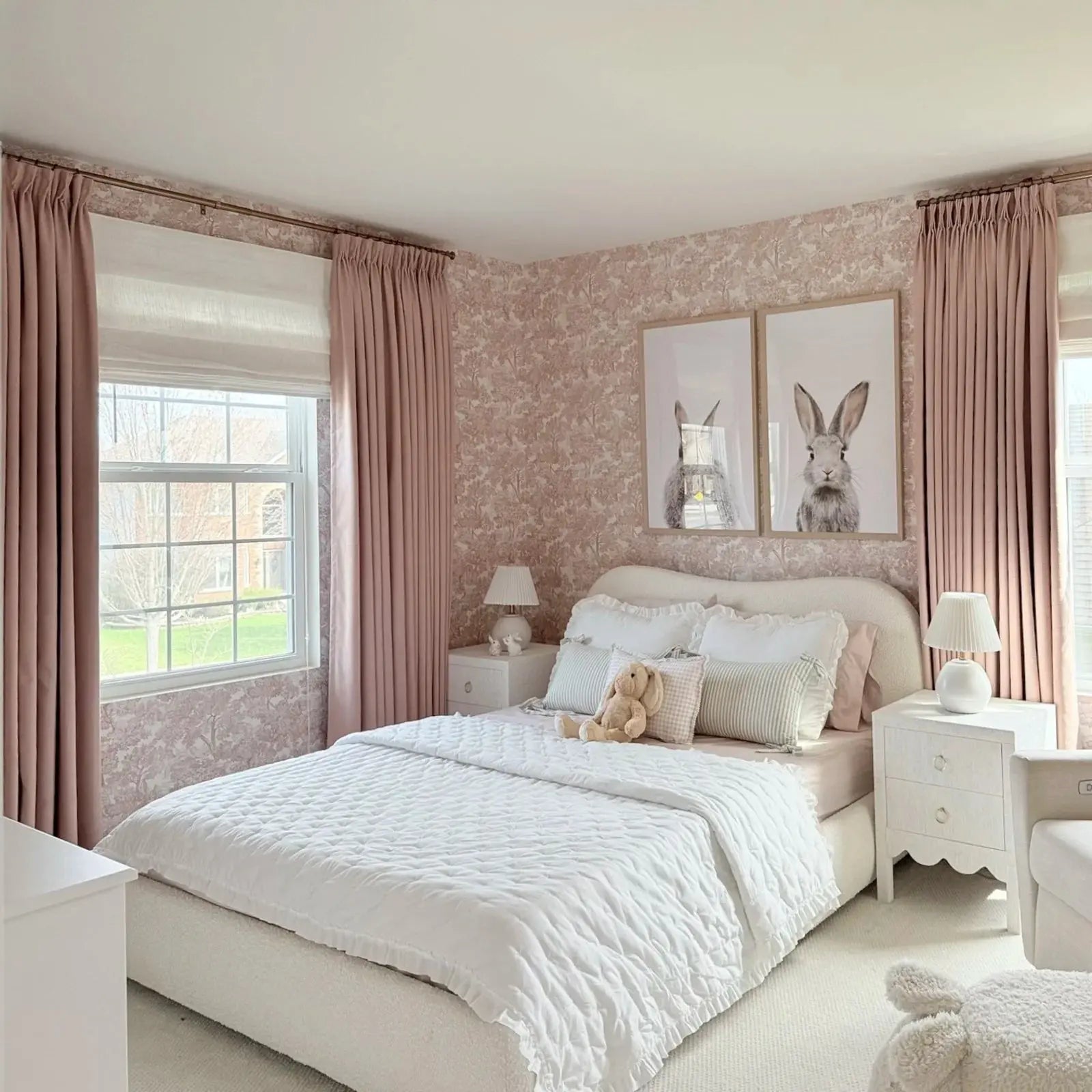 A charming child's bedroom featuring pink toile wallpaper, two large framed black and white bunny portraits, and a white quilted bedspread with a stuffed bunny toy