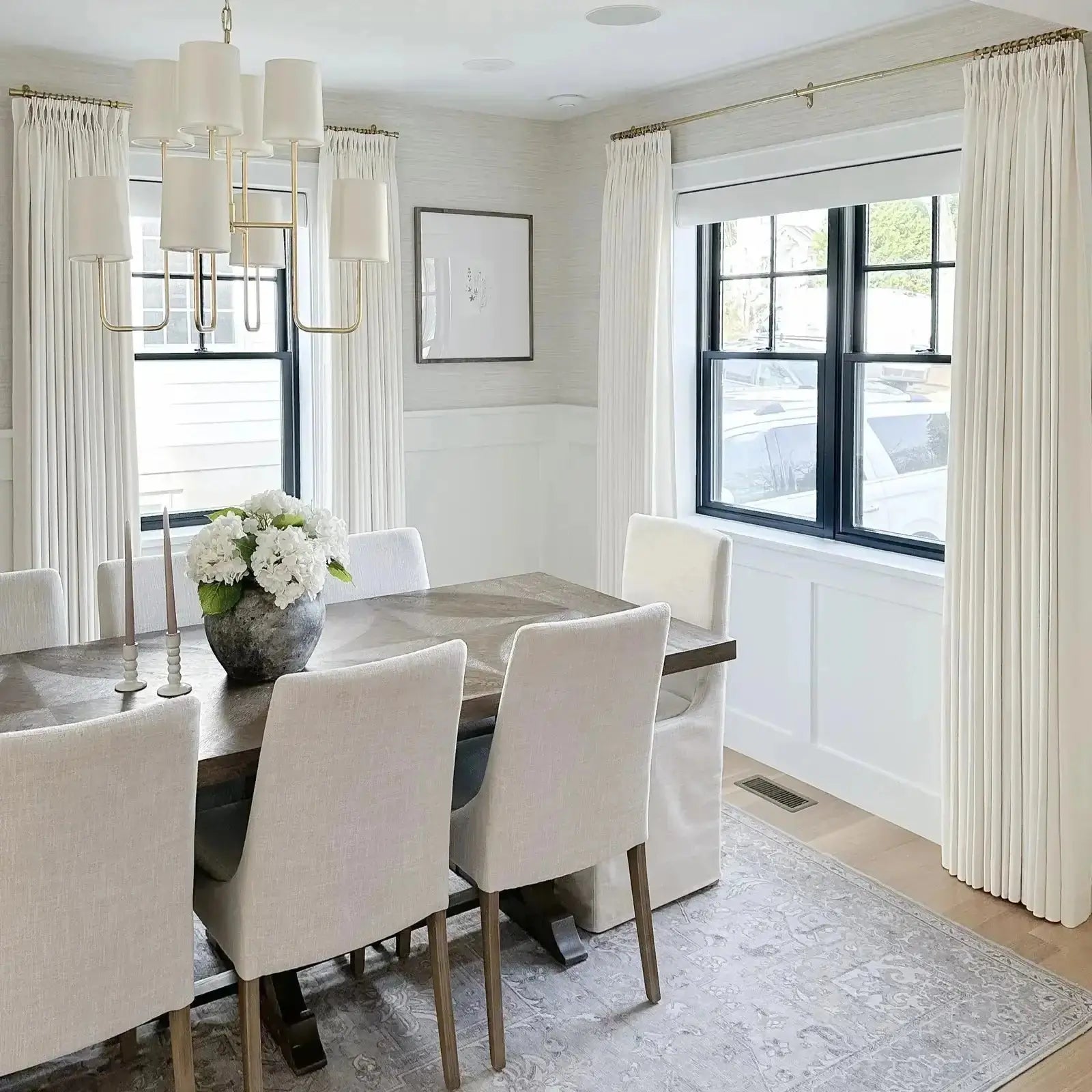 A modern dining room interior with a striking brass chandelier, a dark wood dining table surrounded by light linen chairs, a white hydrangea centerpiece, and cream pleated curtains on brass rods complementing a subtle patterned rug