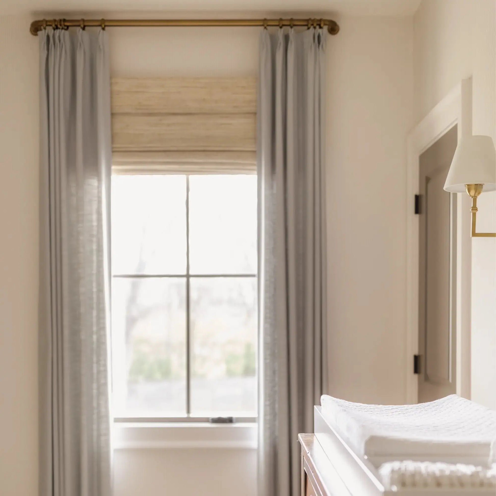 Bright interior of a neutral-toned room showing light gray drapes on a brass rod, a woven Roman shade, and a white changing pad