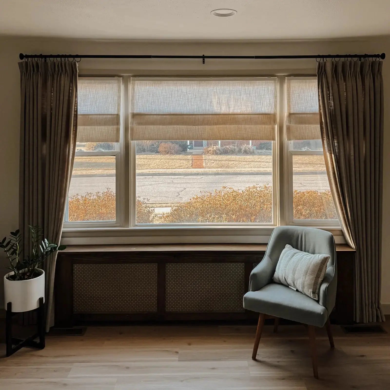 An interior living room features a large three-panel window with beige linen curtains and Roman blinds, a light gray armchair and striped pillow, a dark wood radiator cover with cane webbing, and a potted ZZ plant, overlooking a suburban street and distant house