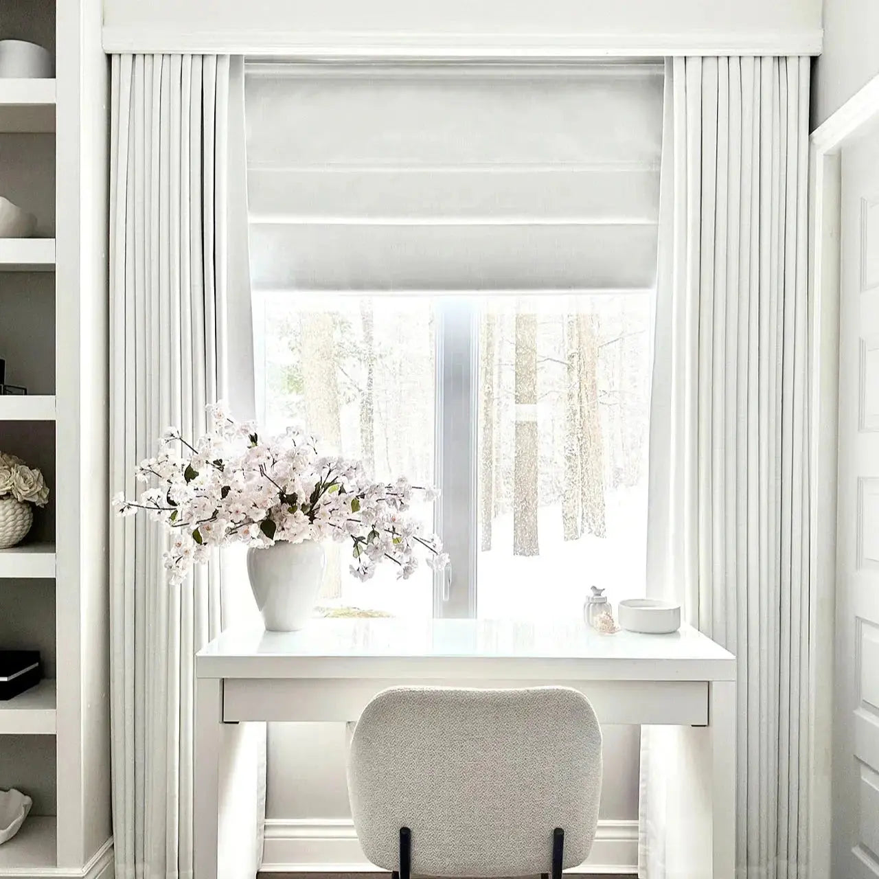 A bright minimalist home office features a white desk with a large white cherry blossom arrangement, a cream textured bouclé chair, and a window revealing a snowy forest outside