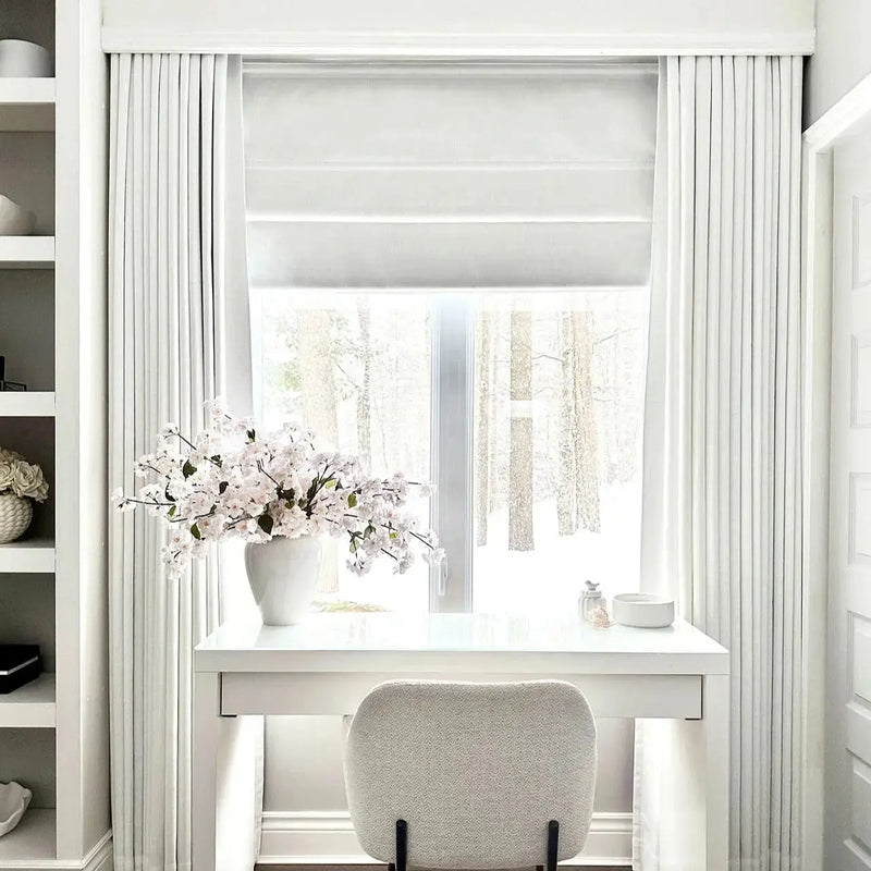 A bright minimalist home office features a white desk with a large white cherry blossom arrangement, a cream textured boucl¨¦ chair, and a window revealing a snowy forest outside