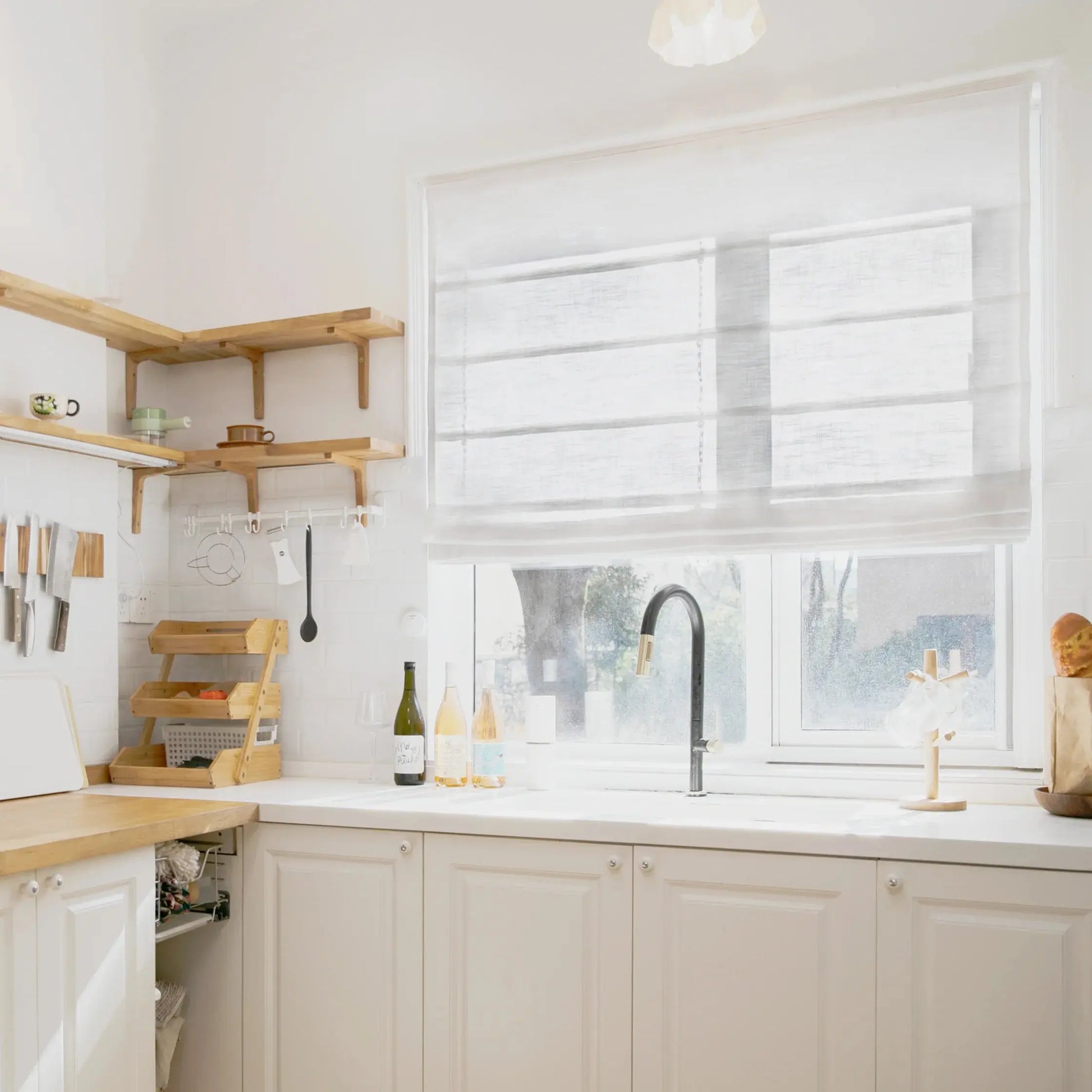 Photograph of a clean modern minimalist kitchen with white shaker cabinets, light wood countertops and open shelving, a prominent black gooseneck faucet, and three decorative wine bottles next to a window with white Roman shades