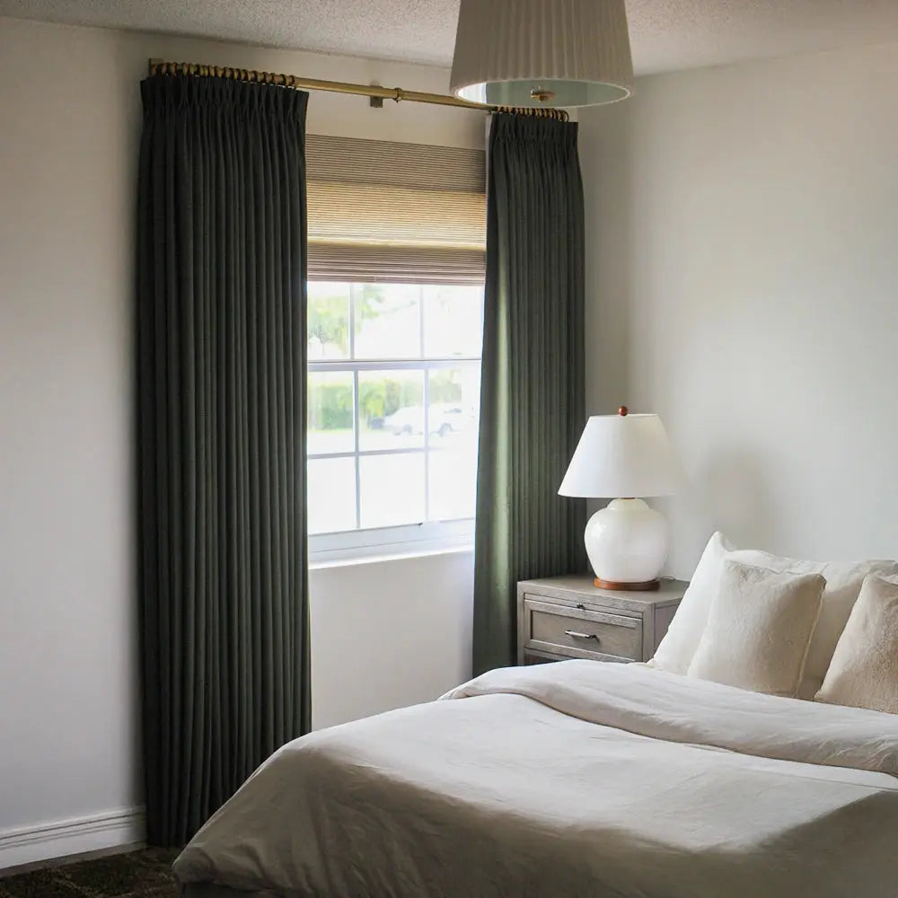 Modern bedroom interior with dark green pleated drapes on a brass rod, a white ceramic table lamp on a gray nightstand, and a light beige bed