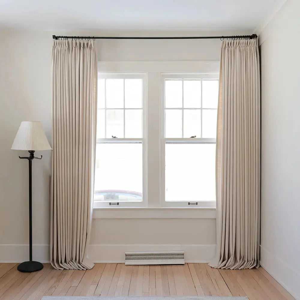 Interior view of a sunlit room featuring two white grid windows flanked by long pleated beige curtains on a black rod, a black floor lamp with a pleated blind, and light hardwood flooring with a white baseboard heater