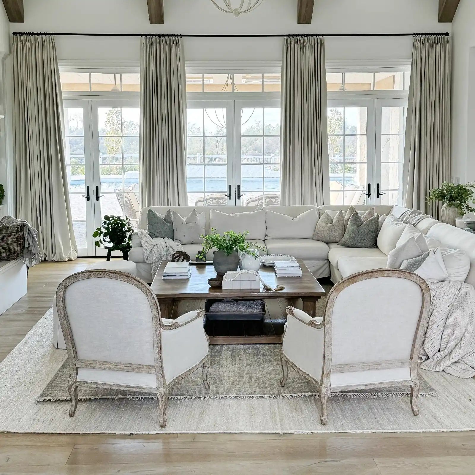 Photograph of a chic indoor living room overlooking an outdoor swimming pool, featuring a large white sectional sofa with neutral throw pillows, a dark wood coffee table adorned with green plants, and two elegant white linen and distressed wood armchairs on a layered rug