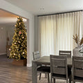 Interior view of a modern dining room featuring a gray wooden table and chairs with dark cushions, looking into a living room with a brightly lit Christmas tree adorned with white, gold, and a unique red handprint ornament, framed by tall off-white curtains and a large gray ceramic vase with dried branches