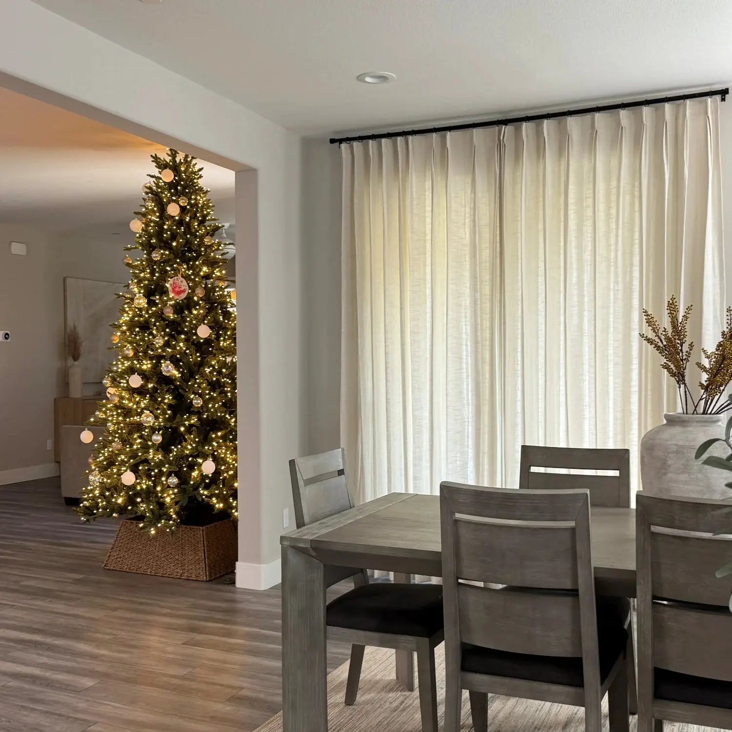 Interior view of a modern dining room featuring a gray wooden table and chairs with dark cushions, looking into a living room with a brightly lit Christmas tree adorned with white, gold, and a unique red handprint ornament, framed by tall off-white curtains and a large gray ceramic vase with dried branches