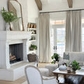 A bright, neutral-toned living room featuring a grand white stone fireplace with dark herringbone tile, an ornate gold mirror, exposed wooden ceiling beams, and French doors opening to a view of a backyard pool, adorned with natural textures and abundant greenery