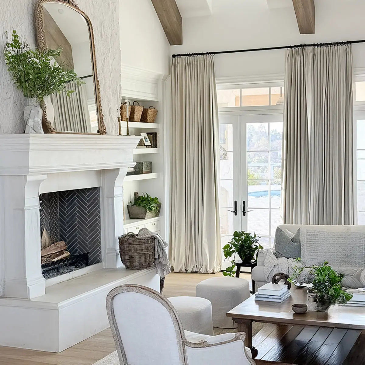 A bright, neutral-toned living room featuring a grand white stone fireplace with dark herringbone tile, an ornate gold mirror, exposed wooden ceiling beams, and French doors opening to a view of a backyard pool, adorned with natural textures and abundant greenery