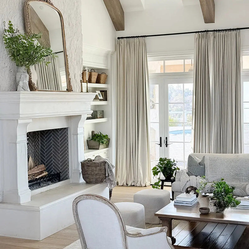 A bright, neutral-toned living room featuring a grand white stone fireplace with dark herringbone tile, an ornate gold mirror, exposed wooden ceiling beams, and French doors opening to a view of a backyard pool, adorned with natural textures and abundant greenery