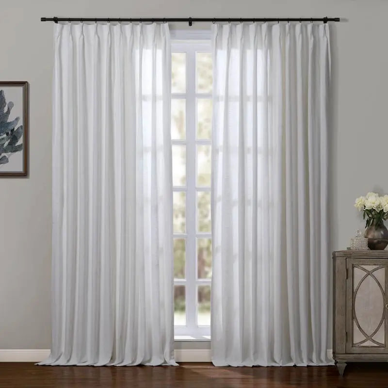 A bright room showcasing sheer white pleated curtains on a black rod flanking a large window with natural light, alongside a distressed wooden cabinet holding a vase of white flowers