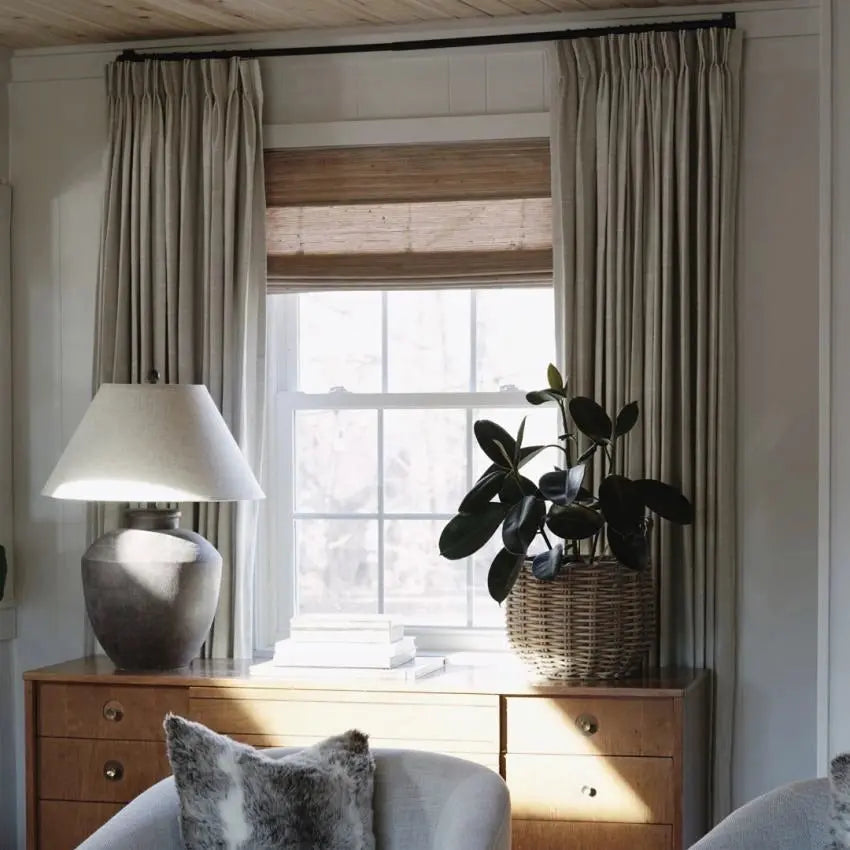 A cozy interior room filled with natural sunlight, featuring a window dressed with light beige drapes and a bamboo blind, a dark green rubber plant in a wicker basket, a rustic ceramic lamp, and a mid-century modern wooden dresser