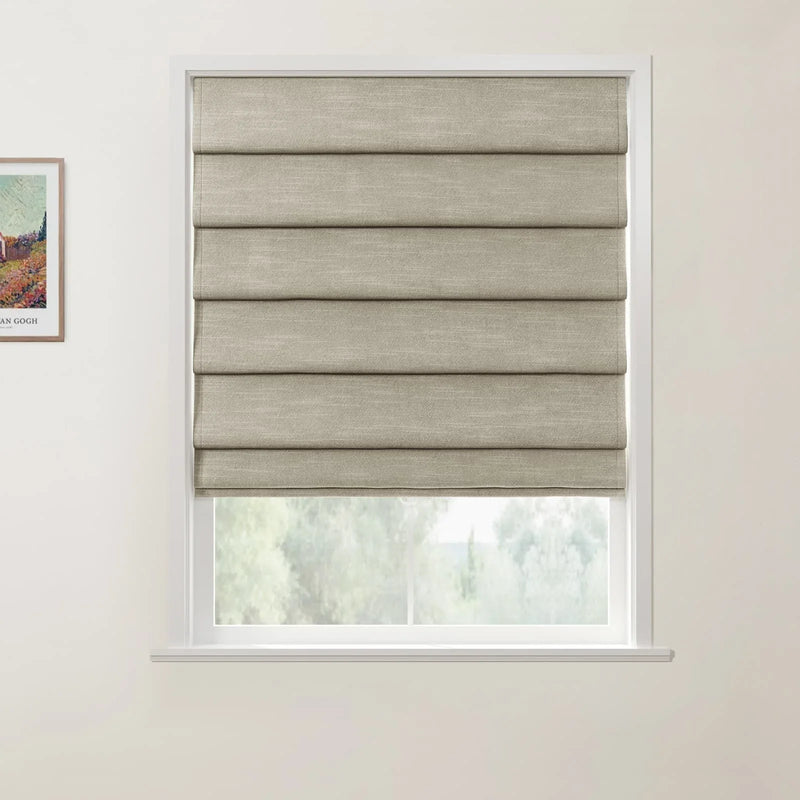 A framed Van Gogh print hangs beside a window fitted with a partially lowered natural beige faux linen Roman blind, revealing blurred green foliage outside