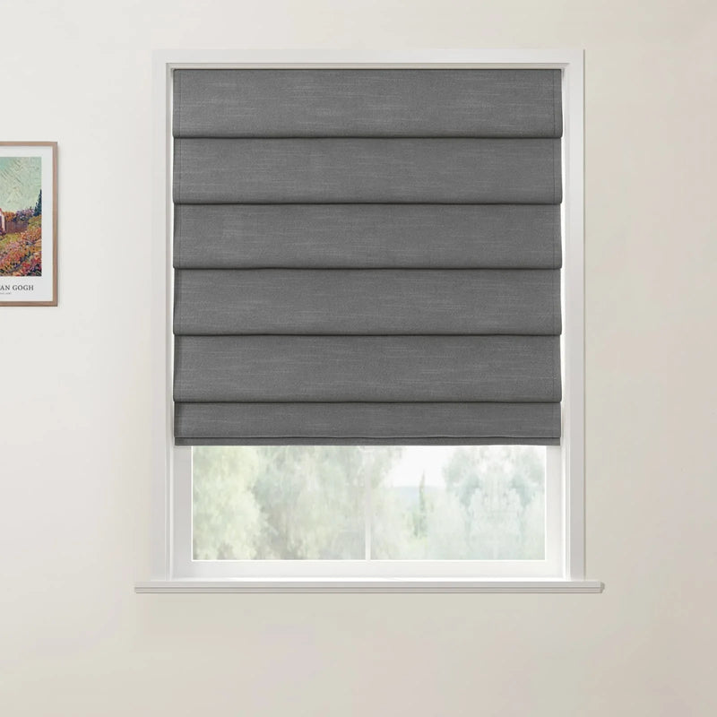 Photograph of a dark grey fabric Roman blind lowered in a white window frame with soft green trees visible outside and a framed Van Gogh print on the left wall