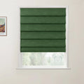 A photograph showcasing a window with a dark green textured fabric Roman blind partially raised above a view of blurred green trees, next to a framed print of a colorful Van Gogh landscape painting on a white wall